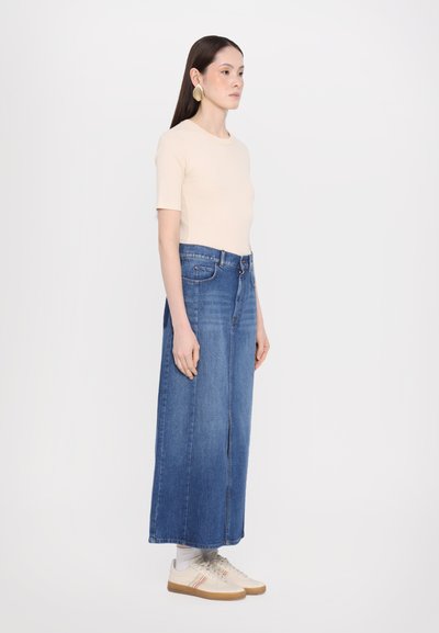 Light beige fitted top, high-waisted long denim skirt, blue wash, side slit, paired with light sneakers featuring colorful stripes.