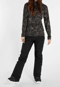 Donna che indossa pantaloni neri e una maglietta nera a maniche lunghe con un motivo a macchie di vernice multicolore, in piedi con le gambe divaricate.
