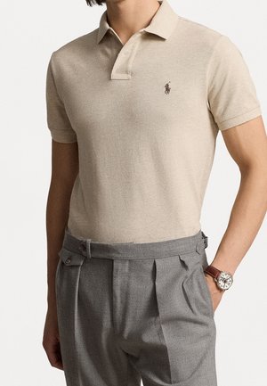 Polo beige de tejido texturizado con tapeta de tres botones, combinado con pantalones gris con pinzas y un reloj de cuero marrón.