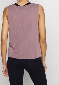 Femme portant un haut sans manches texturé mauve et des leggings noirs, vue de dos sur un fond uni.