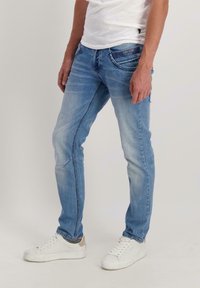 Man in vervaagde blauwe spijkerbroek met een label op de voorkzak en witte sneakers, staand voor een effen lichte achtergrond.