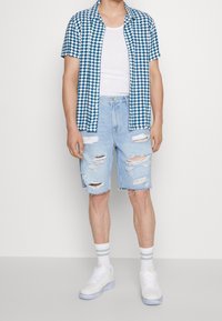 Chemise à manches courtes légère à carreaux bleus et blancs, débardeur blanc, shorts en denim clair déchirés, baskets blanches avec des accents gris.