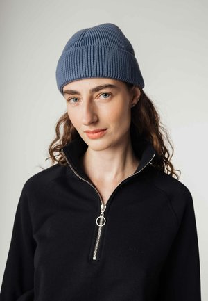 Blauwe geribbelde gebreide beanie met een opgerolde manchet. Model draagt een zwarte ritsvest met een zilveren rits en subtiele branding op de borst.