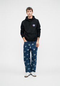 Fiatal férfi fekete Santa Cruz kapucnis pulóverben, kék farmereken fehér yin-yang szimbólumokkal, fehér edzőcipőben és napszemüveggel a fején.