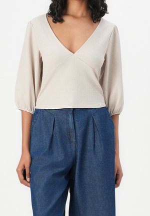 Beige v-kaula-aukollinen pusero, jossa on tekstuurillinen kangas ja pussihihat, yhdistetty korkeavyötäröisiin tummiin farkkuihin, joissa on etutaitteet.