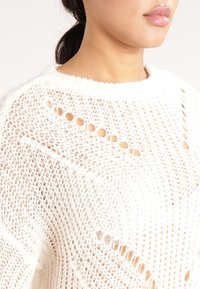 Nainen, jolla on tummat hiukset, valkoisessa, löysästi neulottu villapaita, jossa on tekstuurinen, ilmava kuvio ja pyöreä pääntie.