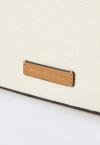 Canvas bag in cream with black trim, featuring a gold metal label embossed with "KARL LAGERFELD" on a tan leather patch.