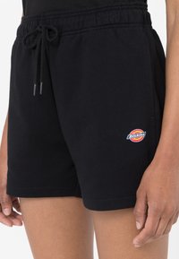 Pantalones cortos de algodón negros con cinturilla elástica y cordón. Presenta un logo colorido de Dickies en la pierna izquierda. Textura suave, diseño casual.