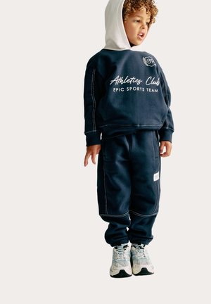 Marineblauwe sportset bestaande uit een hoodie met witte opdruk en bijpassende sweatpants. Inclusief trekkoord en contrasterende stiksels.
