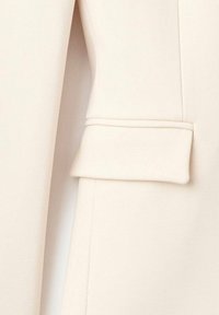 Tela de blazer beige con una textura suave, que presenta un puño doblado, líneas rectas y un diseño minimalista. Vista en primer plano del detalle de la manga.