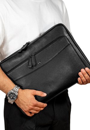 Custodia per laptop in pelle nera con superficie texturizzata, due scomparti con zip e un piccolo logo. Tenuta tra le mani su uno sfondo bianco.