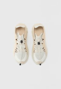 Chaussures à lacets beige et blanc avec panneaux en maille, semelles texturées et bascules noires pour un ajustement réglable ; elles présentent un design épuré.
