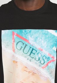 Camiseta negra con un estampado colorido que presenta agua turquesa, playa de arena y un logotipo triangular rojo con la palabra "GUESS" en negro.