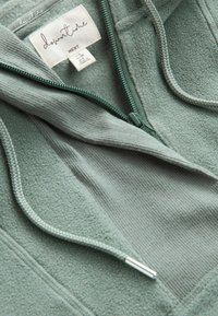 Grøn hættejakke med lynlås i fleece, der har en blød tekstur, kontrasterende ribbet stof og matchende snore med sølvfarvet hardware.