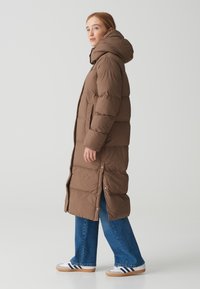 Brauner Puffer-Mantel mit Kapuze, mit einem gesteppte Design, seitlichen Knöpfen und einer langen Silhouette, kombiniert mit blauen Jeans im Schlag.