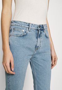 Jean en denim bleu clair à taille haute, coupe droite, cinq poches, et garnitures ton argenté. Le tissu présente une texture subtile.