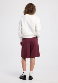 Valkoinen huppari, jossa on ribbisaumat reunuksissa, yhdistettynä burgundinvärisiin laskoshousuihin. Mustat kengät ja sydänkuvioiset valkoiset sukat ovat käytössä.