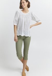 Blouse blanche à manches bouffantes et ourlet brodé, associée à un pantalon court olive. Le modèle porte des ballerines beige clair.