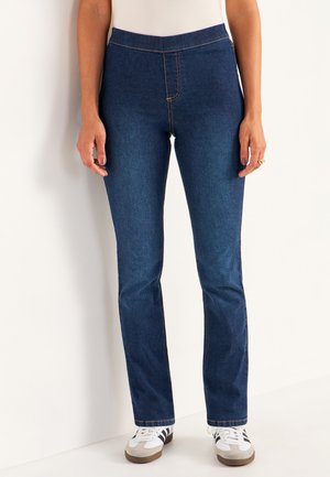 Vrouw die donkerblauwe slim-fit jeans met contrasterende stiksels draagt, witte sneakers met zwarte strepen, staand tegen een witte muur.