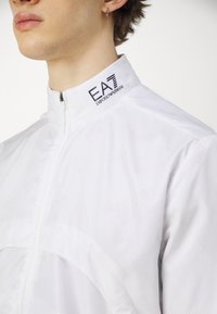 Bela zip-up jakna z visokim ovratnikom, s teksturiranim vzorcem in vtisnjenim logotipom "EA7 Emporio Armani" na ovratniku. Lahka material.