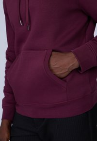 Sweatshirt bordeaux avec une poche frontale, des poignets et un ourlet côtelés, présentant une texture lisse et un cordon de serrage réglable pour la capuche.