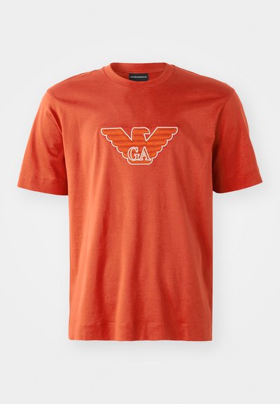 Raudona medvilninė marškinėliai su ryškiu erelio logotipu ir balta bei oranžine siuvinėjimu "GA" ant krūtinės. Trumpomis rankovėmis, apvaliu kaklu.