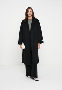 Black wool trench coat with wide lapels, long sleeves, and side pockets. Model wears gray top and black pants, holding a small white bag.
