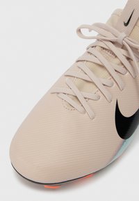 Ανοιχτό μπεζ ποδοσφαιρικό παπούτσι Nike με αντίστοιχους κορδόνες, μαύρο λογότυπο swoosh και μικρούς πορτοκαλί τάκους ορατούς στην άκρη της σόλας.