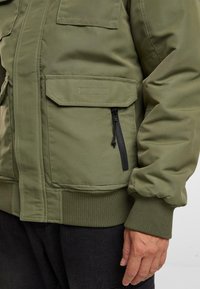 Chaqueta aislante de color verde olivo con puños acanalados, bolsillo con cremallera y dos bolsillos de parche, que presenta una tela suave y un diseño de corte relajado.