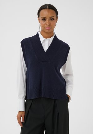 Femme portant un gilet sans manches bleu marine par-dessus une chemise blanche boutonnée et un pantalon noir, debout avec une main dans la poche.