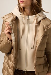 Beige puffer jacket with a smooth, shiny exterior and a soft, hooded sweatshirt underneath. Features gold-tone zippers and drawstrings.