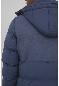 Navy puffer jacket with a textured fabric panel on the upper back and a hood. Features quilted sections and a discreet logo label.