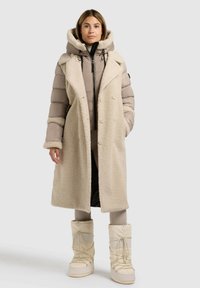 Beidzot bēšā samta garā mēteli ar lielu apkakli, kārtota virs pelēka pūkaina jakas. Kombinēts ar krēmkrāsas līdz ceļiem augstiem zābakiem un pieguļošām biksēm.