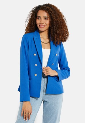 Blauw dubbelzijdig colbert met gouden knopen, ingeknipte revers en een getailleerde pasvorm. Draagt over een witte top met lichte denim jeans.