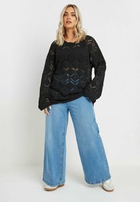 Svart stickad tröja med diamantmönster, vida ärmar och avslappnad passform, parat med lösa blå denimjeans och ljusa sneakers.