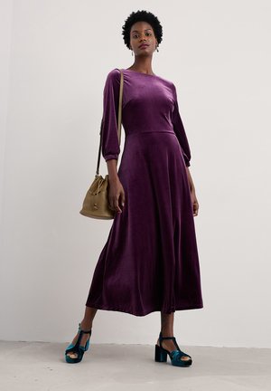 Lila Samtkleid mit langen, gepufften Ärmeln und einer ausgestellten Silhouette, kombiniert mit türkisfarbenen Schuhen und einer beigen Bucket Bag.