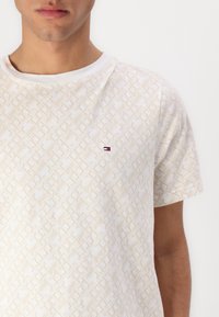 Balts īsām piedurkņu t-krekls ar teksturētu ģeometrisku rakstu, piedāvājot nelielu izšūtu logo sarkanā, baltā un zilā krāsā uz krūtīm.