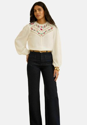 Kobieta w kremowej bluzce z kwiatowym haftem i koronkowymi detalami, dobrana do czarnych, szerokich spodni z wysokim stanem i brązowego paska.