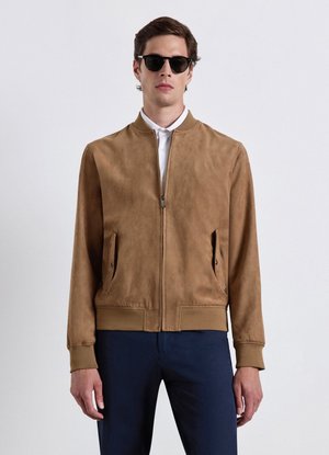 Uomo con occhiali da sole neri, giacca marrone con zip e tasche, camicia bianca e pantaloni blu scuro, in piedi davanti a uno sfondo chiaro semplice.