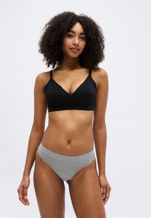 Femme aux cheveux bouclés portant un soutien-gorge noir et un sous-vêtement gris, debout devant un fond uni clair.