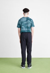 Mann mit kurzem, lockigem Haar steht mit dem Rücken zur Kamera, trägt ein türkisfarbenes Hemd mit abstraktem Muster, schwarze Hosen und schwarze Air Jordan Sneakers auf einem grünen Boden.