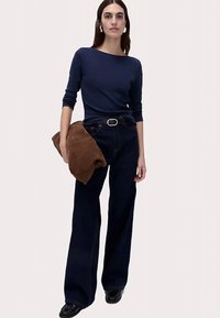 Granatowa bluzka z długim rękawem w połączeniu z ciemnoniebieskimi spodniami o szerokich nogawkach. Modelka trzyma brązową saszetkę, nosi czarne buty i duże białe kolczyki.