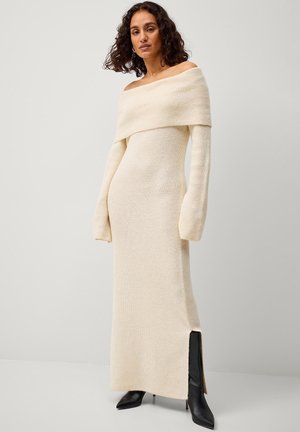 Cremefarbenes Strickkleid mit weitem Ausschnitt, langen Ärmeln und seitlichem Schlitz; strukturierter Stoff; figurbetonte Silhouette. Schwarze Absatzstiefel getragen.