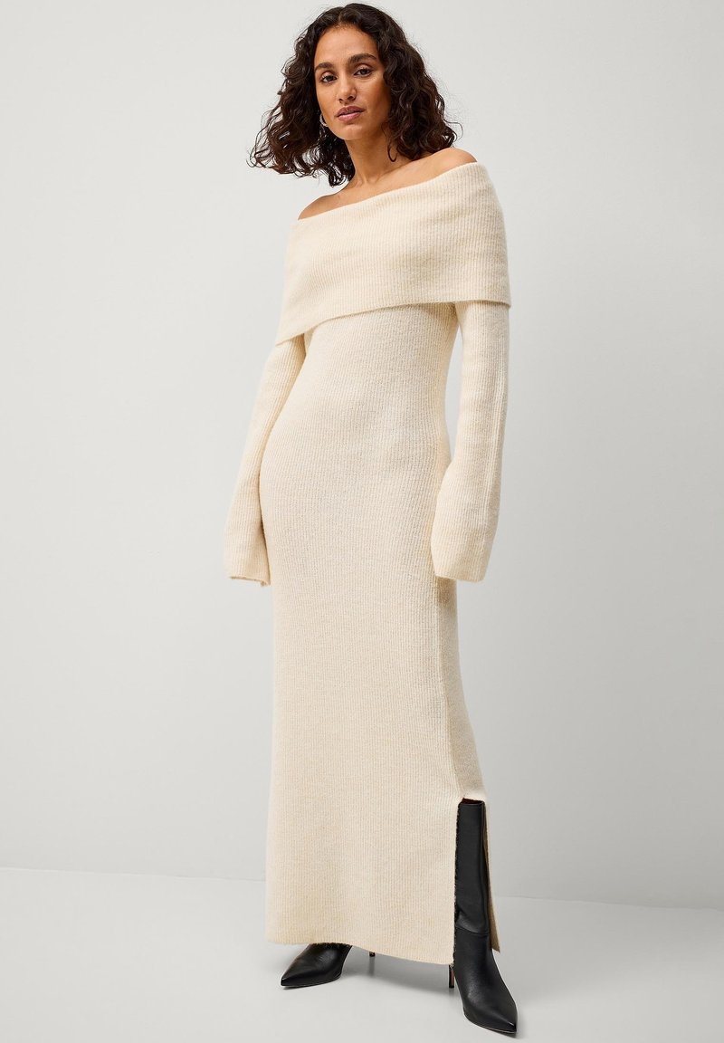 Off-shoulder, crèmekleurige gebreide jurk met lange mouwen en een zijsplit; gestructureerde stof; aansluitende pasvorm. Zwarte gehandhaakte laarzen dragen.