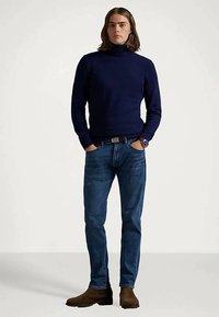 Νέος άνδρας που στέκεται με τα χέρια στις τσέπες, φορώντας ένα ναυτικό μπλε πουλόβερ με ζιβάγκο, μπλε τζιν, καφέ μπότες και ρολόι στον αριστερό καρπό.