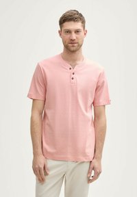 Ung mand iført en lyserød kortærmet henley skjorte med tre knapper og lysebeige bukser, stående mod en ensfarvet lyse baggrund.