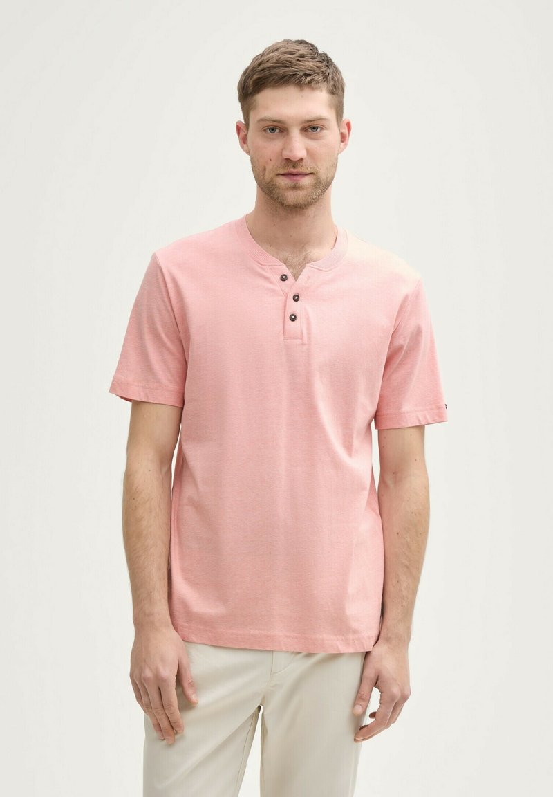 Ung mand iført en lyserød kortærmet henley skjorte med tre knapper og lysebeige bukser, stående mod en ensfarvet lyse baggrund.