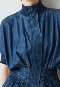 Denimklänning med hög, ribbad krage, framfåror och samlad midja. Djupblå färg med en slät yta och subtil lyster.