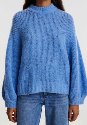 Kvinde iført en løs, struktureret lyseblå striktrøje med ballonærmer, parret med medium vaskede blå jeans.