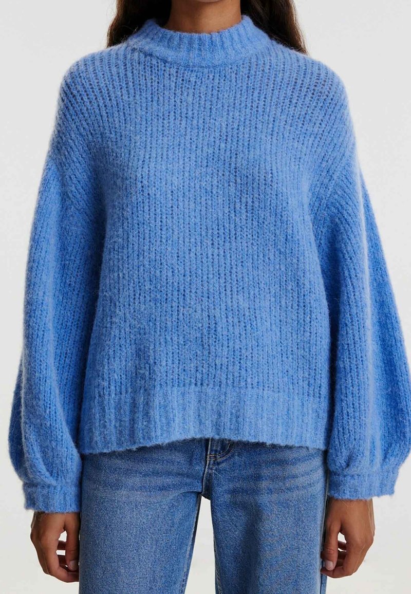 Femme portant un pull en tricot léger bleu clair, ample et texturé, avec des manches ballons, associé à un jean bleu de teinte moyenne.